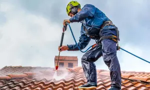 Pracownik w kasku i zabezpieczeniach myje dach myjką ciśnieniową. Sprawdź, jaka jest mycie dachu cena i odzyskaj blask swojej posesji.