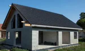 Dom w stanie surowym otwartym, z dachem pokrytym czarną dachówką. Widać otwory okienne i drzwiowe. Idealny do dalszych prac budowlanych.