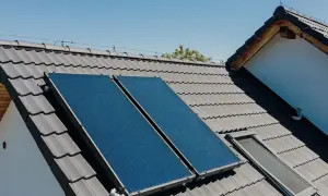 Dwa panele instalacji solarnej na szarym dachu, obok okna dachowego. Słońce ogrzewa dom.