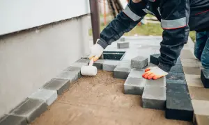 Pracownik układa kostkę brukową, tworząc opaskę wokół domu. Widać jego ręce w rękawicach i gumowy młotek.