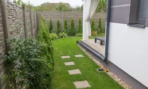 Nowoczesny ogród z kamiennym murem i zadbanym trawnikiem. Rynna odprowadza wodę z dachu na trawnik, gdzie znajduje się ścieżka z płyt.