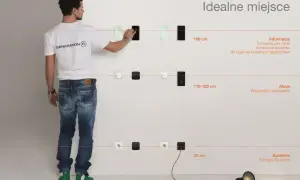 Mężczyzna pokazuje na ścianie, na jakiej wysokości gniazdka są idealne: 150 cm dla informacji, 110-120 cm dla akcji, 30 cm dla zasilania.