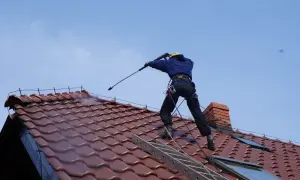 Pracownik w kasku i zabezpieczeniach wykonuje mycie dachu z użyciem myjki ciśnieniowej.