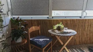 Przytulny balkon typu loggia z drewnianym stolikiem, krzesłem i roślinami. Rolety częściowo zasłonięte.