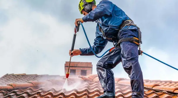 Pracownik w kasku i zabezpieczeniach myje dach myjką ciśnieniową. Sprawdź, jaka jest mycie dachu cena i odzyskaj blask swojej posesji.