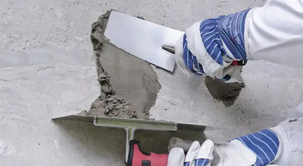 Naprawa betonu na zewnątrz: ręce w rękawicach nakładają zaprawę kielnią na uszkodzony fragment ściany.