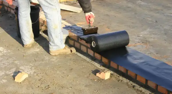 Pracownik kładzie hydroizolację na murze, przygotowując zbrojenie pod dalsze prace budowlane.