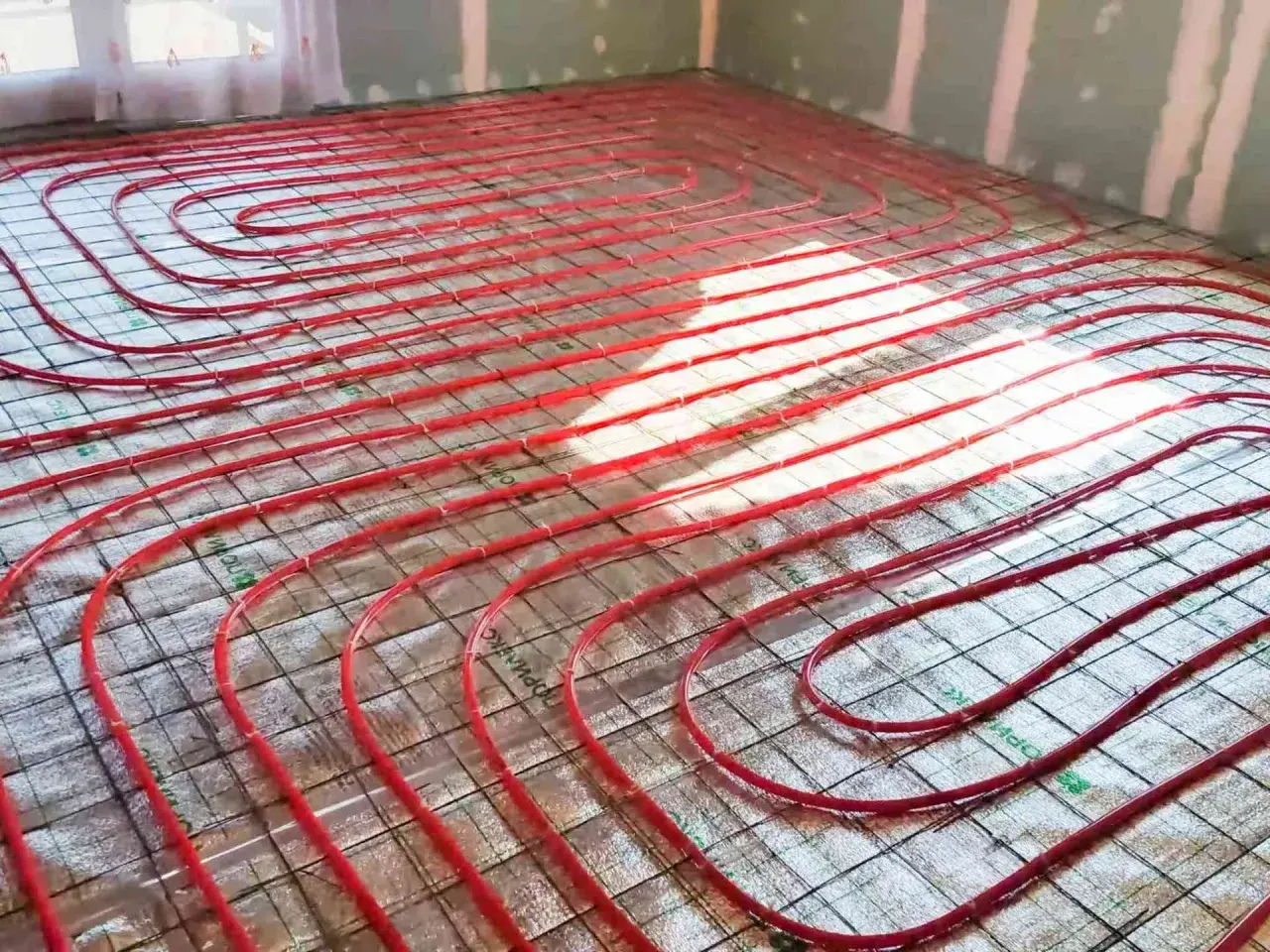 Instalacja ogrzewania podłogowego. Czerwone rurki ułożone na siatce, przygotowane do wylania betonu. To inwestycja, która obniży koszt ogrzewania.