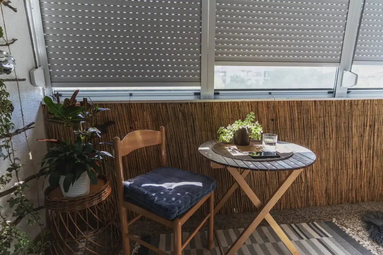 Przytulny balkon typu loggia z drewnianym stolikiem, krzesłem i roślinami. Rolety częściowo zasłonięte.