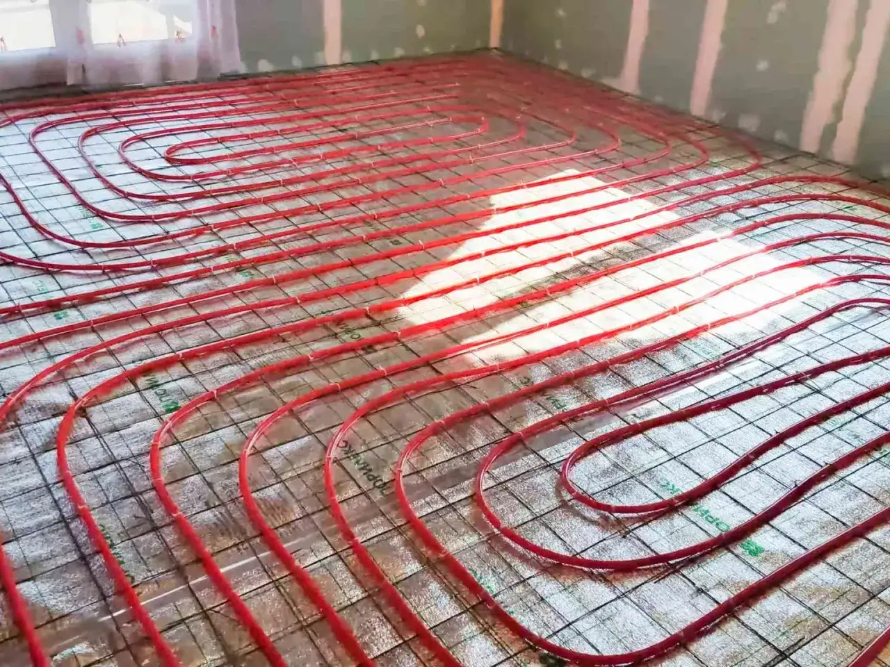 Instalacja ogrzewania podłogowego. Czerwone rurki ułożone na siatce, przygotowane do wylania betonu. To inwestycja, która obniży koszt ogrzewania.