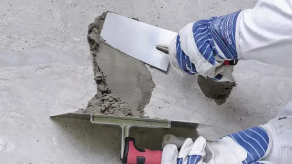 Naprawa betonu na zewnątrz: ręce w rękawicach nakładają zaprawę kielnią na uszkodzony fragment ściany.