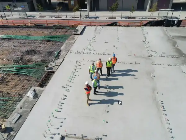 Grupa budowlańców na placu budowy, dyskutująca o cenie betonu i postępach prac. W tle zbrojenie i świeżo wylany beton.