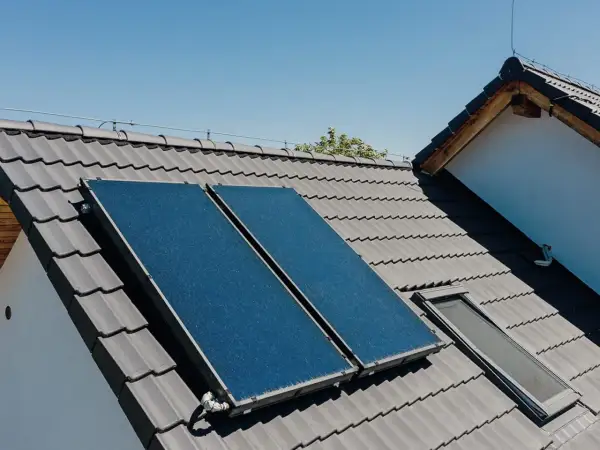 Dwa panele instalacji solarnej na szarym dachu, obok okna dachowego. Słońce ogrzewa dom.