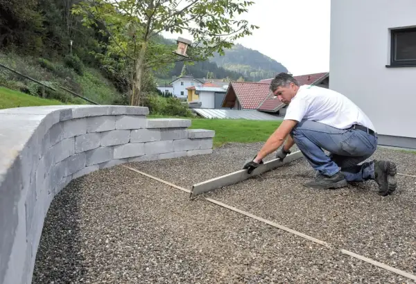 Mężczyzna wyrównuje podjazd z betonu szczotkowanego. Obok wznosi się mur z szarego kamienia.