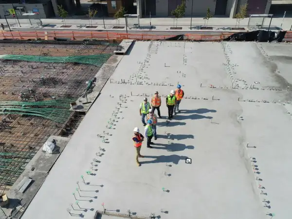 Grupa budowlańców na placu budowy, dyskutująca o kosztach kubika betonu. W tle zbrojenie i wylana płyta.