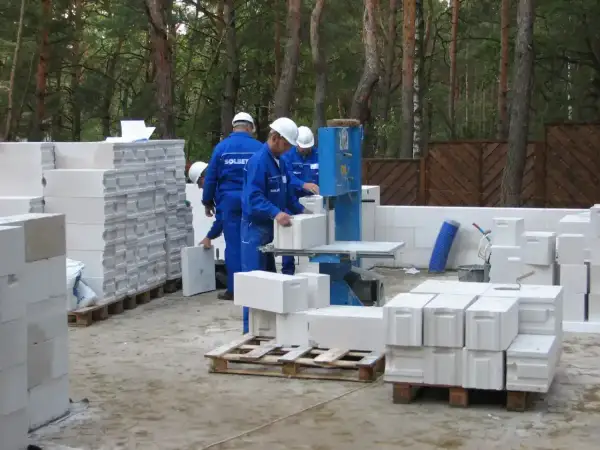 Budowa domu z bloczków betonowych. Pracownicy w niebieskich kombinezonach i białych kaskach układają materiał. Widać piłę do cięcia bloczków.