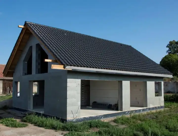 Dom w stanie surowym otwartym, z dachem pokrytym czarną dachówką. Widać otwory okienne i drzwiowe. Idealny do dalszych prac budowlanych.